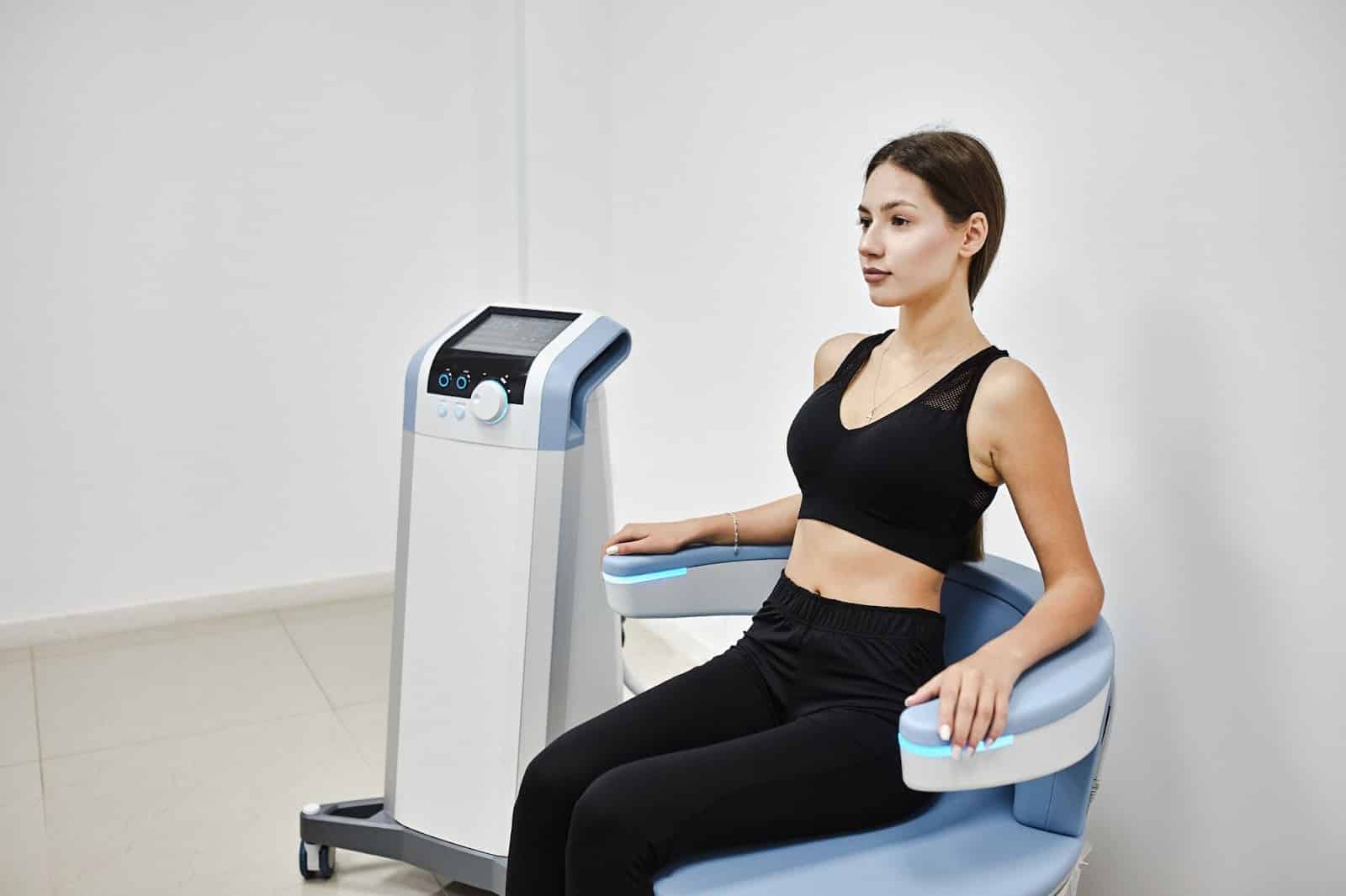 A woman seated on an Emsella chair, designed for incontinence treatment, in front of a therapeutic machine.